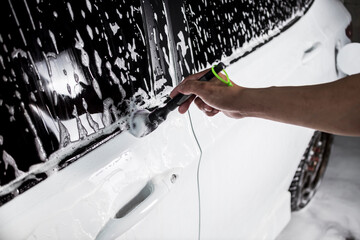 Wall Mural - A man cleans the outer window seal of a white sedan covered in car shampoo with a fine brush. A vehicle being serviced at a carwash or auto detailing shop.