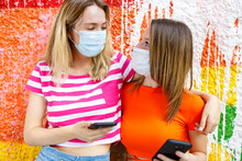 Female Friends In Protective Face Masks Looking At Each Other While Arms Around Against Colorful Wall