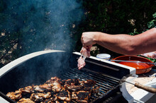 Hand Of Young Man Placing Meat On Barbecue Grill In Yard