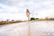 Cheerful Girl Splashing Water In Puddle While Holding Umbrella Against Cloudy Sky