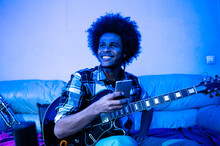 Happy Afro Man With Guitar Looking Away While Holding Mobile Phone In Blue Room