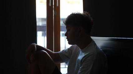 Depressed Asian man sitting by the window in bedroom with stressed and hopeless face. Loneliness sad man being alone in bedroom thoughtful with negative emotion. Mental health and life problem concept