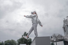Girl In Silver Suit Looking Through VR Goggles, Standing On Box, Against Grey Sky