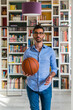 © Giorgio Magini/Westend61 - Portrait of young man with basketball standing in front of bookshelves at home
