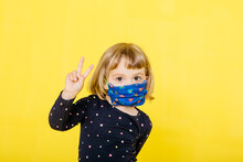 Portrait Of Cute Blond Girl Wearing Face Mask And Gesturing Peace Sign Against Yellow Background