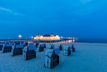 Germany, Mecklenburg-Western Pomerania, Heringsdorf, Hooded Beach Chairs On Sandy Coastal Beach At Dusk With Illuminated Pier In Background