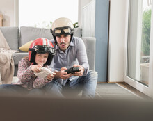Young Man And Little Girl Wearing Biker Helmets, Playing Racing Game With Gaming Consoles