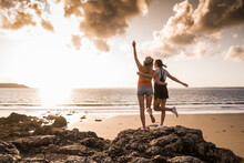 Two Girlfriends Standing On Rocky Beach, Waving At Sunset, Rear View