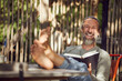 © Maya Claussen/Westend61 - Cheerful bearded man reading book while relaxing on chair in yard