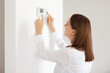 © sementsova321 - Good looking young adult woman wearing white casual style shirt, using connected appliances device on wall, monitoring energy security heating system, digital control in apartment.