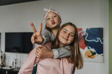Smiling Girl Piggybacking Friend Showing Victory Sign While Standing At Home