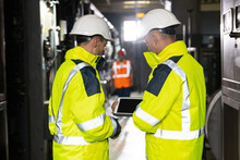 Two Heavy Industry Engineers Stand In Pipe Manufacturing Factory, Use Digital Tablet Computer, Have Discussion. Facility For Construction Of Oil, Gas And Fuel Pipeline Transportation Products.