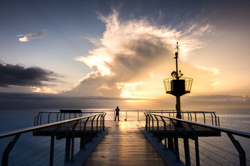 Wall Mural - Person standing on the bridge over the calm sea under a cloudy sky at sunset in Spain