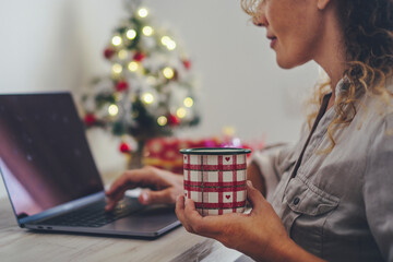 Wall Mural - Close up of christmas mug with tea or coffee and happy woman working alone on computer laptop at home. Young adult female people in holidays december season using internet