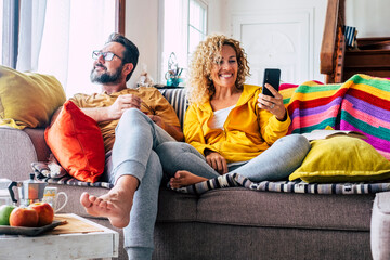 Happy adult couple man and woman enjoy time at home together in morning breakfast. Relationship lifestyle people in indoor leisure activity using phone and looking outside the window