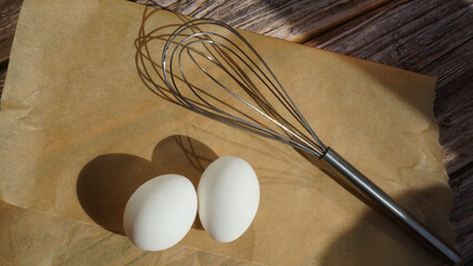 two white chicken eggs metal whisk for whipping on parchment paper on a wooden surface sun rays breakfast