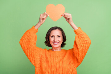 Poster - Photo of cheerful elderly woman hold paper postcard heart shape valentine day isolated over green color background