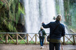 © AliciaFdez - Anciano con un palo selfie tomando una foto a su esposa con su teléfono móvil frente a una enorme cascada en un parque natural
