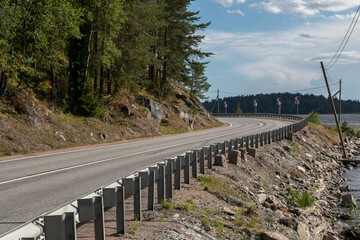 Curve of an empty asphalt countryside highway. The road has 2 lanes, fenced and with white markings. Large stones, rocks and trees is on the side of the road, on the other side there is a Ladoga lake.