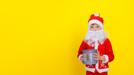 Medium portrait of a cute child boy in Santa Claus clothes and with an artificial beard, who is holding a round gift box with the words New Year 2022, yellow, copy space.