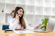 © kamiphotos - Beautiful Asian businesswoman sitting in her room, talking to her partner via mobile phone and checking financial documents, she is a female executive of a startup company. Financial management.