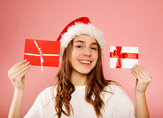 Happy caucasian girl teenager standing in white t-shirt and santa claus red hat holding two gift certificates or greeting inviting cards over pink background. Studio portrait. Christmas and New year 