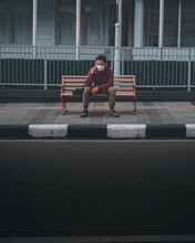 Young Indonesian Man With A Face Mask Talking On The Phone While Sitting Alone On The Bench
