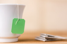Tea Bag With A Green Label In A Ceramic White Cup On A Table Top, Warm Yellow Background, Closeup View Of Mock-up Template