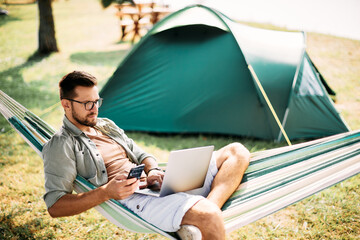 Wall Mural - mid adult man uses smart phone and laptop while relaxing in hammock in nature.