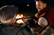 © Drazen - Happy man plays acoustic guitar while relaxing by campfire with is mature father at night.