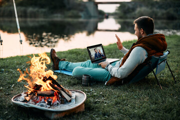 Wall Mural - happy man has video call with business co-worker while relaxing by campfire and fishing in nature.