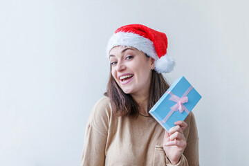 Wall Mural - Beautiful girl in red Santa Claus hat holding blue gift box in hand isolated on white background. Young woman portrait, true emotions. Happy Christmas and New Year holidays concept.