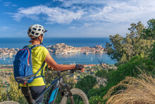 Nice Woman Riding Her Electric Mountain Bike At The Coastline Of Mediterranean Sea On The Island Of Elba In The Tuscan Archipelago, In Front Of Porto Ferraio,Tuscany, Italy