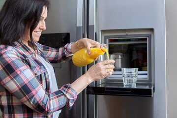 Wall Mural - Middle aged woman pouring orange juice into a glass from the refrigerator