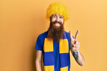 Sticker - Redhead man with long beard football hooligan cheering game wearing funny wig showing and pointing up with fingers number two while smiling confident and happy.