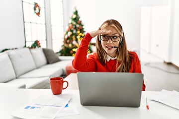 Poster - Young caucasian girl sitting on the table working using laptop by christmas tree very happy and smiling looking far away with hand over head. searching concept.