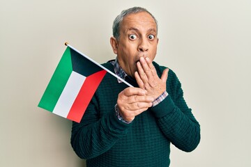 Canvas Print - Handsome senior man with grey hair holding kuwait flag covering mouth with hand, shocked and afraid for mistake. surprised expression