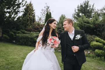 A fashionable groom in a black suit and a beautiful brunette bride in a white dress with a bouquet in her hands are walking in the park holding hands. Wedding portrait of smiling newlyweds.