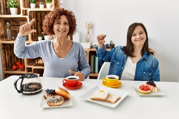 Poster - Family of mother and down syndrome daughter sitting at home eating breakfast smiling and confident gesturing with hand doing small size sign with fingers looking and the camera. measure concept.