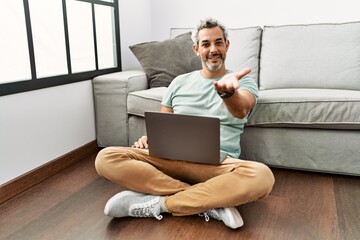 Canvas Print - Middle age hispanic man using laptop sitting on the floor at the living room smiling cheerful offering palm hand giving assistance and acceptance.