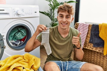 Sticker - Young caucasian man doing laundry holding sock smiling happy and positive, thumb up doing excellent and approval sign