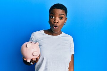 Sticker - Young african american woman holding piggy bank scared and amazed with open mouth for surprise, disbelief face