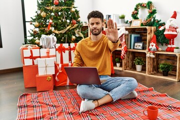 Sticker - Arab young man using laptop sitting by christmas tree doing stop sing with palm of the hand. warning expression with negative and serious gesture on the face.