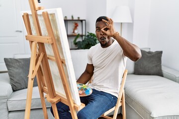 Wall Mural - Young african man painting on canvas at home doing ok gesture shocked with surprised face, eye looking through fingers. unbelieving expression.