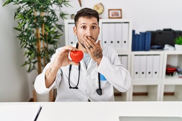 Wall Mural - Young doctor holding heart at medical clinic covering mouth with hand, shocked and afraid for mistake. surprised expression