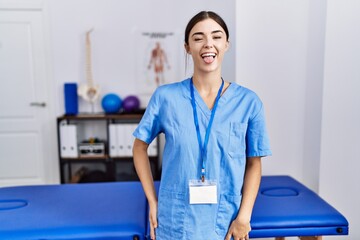 Poster - Young hispanic woman wearing physiotherapist uniform standing at clinic sticking tongue out happy with funny expression. emotion concept.