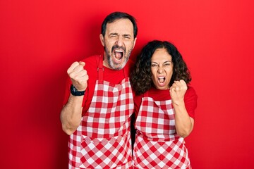 Wall Mural - Middle age couple of hispanic woman and man wearing cook apron angry and mad raising fist frustrated and furious while shouting with anger. rage and aggressive concept.