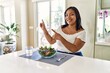 © Krakenimages.com - Young hispanic woman eating healthy salad at home smiling and looking at the camera pointing with two hands and fingers to the side.