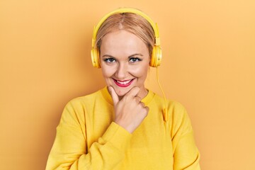 Canvas Print - Young blonde woman listening to music using headphones looking confident at the camera smiling with crossed arms and hand raised on chin. thinking positive.