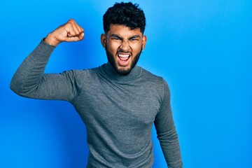 Canvas Print - Arab man with beard wearing turtleneck sweater angry and mad raising fist frustrated and furious while shouting with anger. rage and aggressive concept.